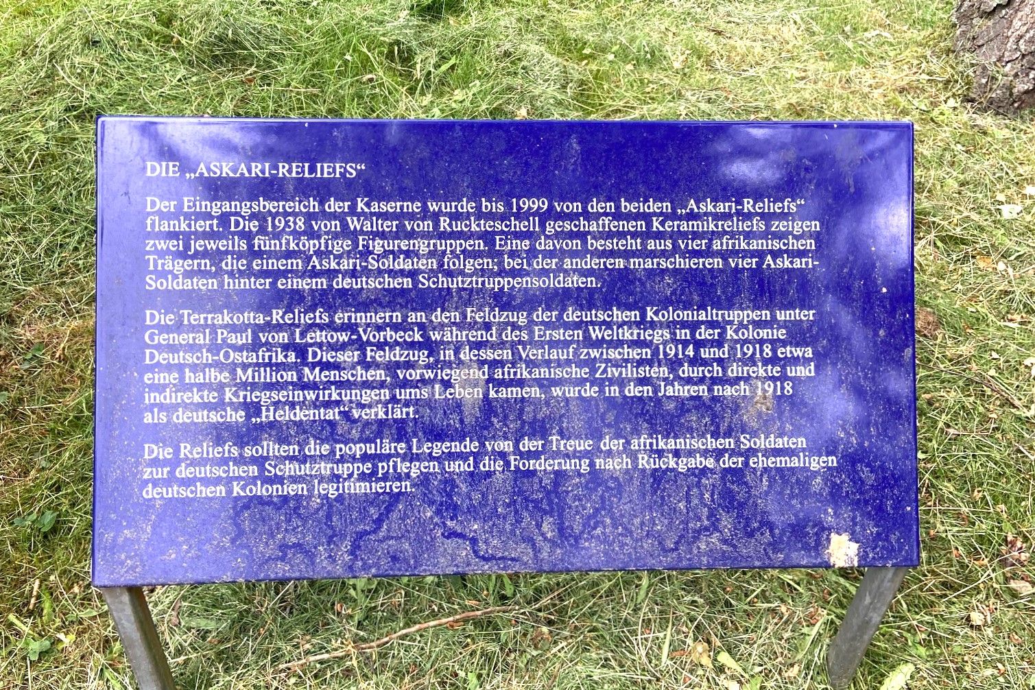 Bild von einer blauen Infotafel: DIE „ASKARI-RELIEFS" Der Eingangsbereich der Kaserne wurde bis 1999 von den beiden „Askari-Reliefs" flankiert. Die 1938 von Walter von Ruckteschell geschaffenen Keramikreliefs zeigen zwei jeweils fünfköpfige Figurengruppen. Eine davon besteht aus vier afrikanischen Trägern, die einem Askari-Soldaten folgen; bei der anderen marschieren vier Askari-Soldaten hinter einem deutschen Schutztruppensoldaten. Die Terrakotta-Reliefs erinnern an den Feldzug der deutschen Kolonialtruppen unter General Paul von Lettow-Vorbeck während des Ersten Weltkriegs in der Kolonie Deutsch-Ostafrika. Dieser Feldzug, in dessen Verlauf zwischen 1914 und 1918 etwa eine halbe Million Menschen, vorwiegend afrikanische Zivilisten, durch direkte und indirekte Kriegseinwirkungen ums Leben kamen, wurde in den Jahren nach 1918 als deutsche „Heldentat" verklärt. Die Reliefs sollten die populäre Legende von der Treue der afrikanischen Soldaten zur deutschen Schutztruppe pflegen und die Forderung nach Rückgabe der ehemaligen deutschen Kolonien legitimieren.